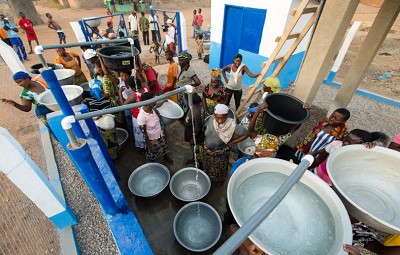 People gathering and filling large metal water buckets from several faucets - Nyani Quarmyne/ Panos